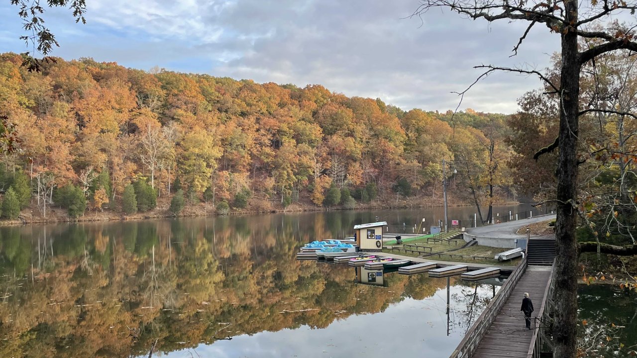 Autumn scene with a calm lake reflecting vibrant orange and green trees. A lone person walks on a wooden boardwalk by the water.