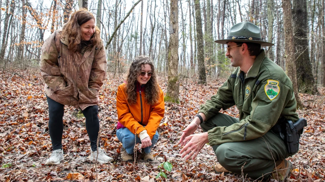 Park ranger and two women crouching in a forest, smiling and observing the ground.