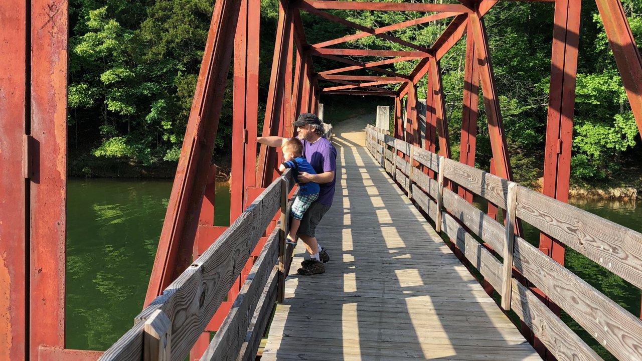 A person in a purple shirt and cap stands with a child on a sunlit, red metal truss bridge over a river, surrounded by lush green trees, conveying a peaceful, scenic moment.