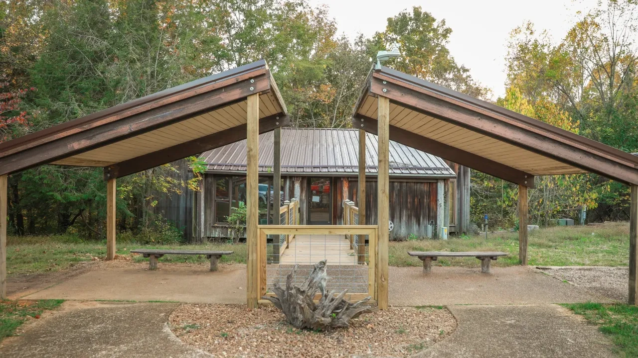 Wooden pavilion leading to rustic building with trees in the background.