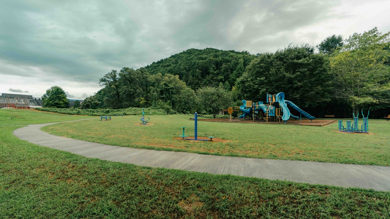 view of playground with a concrete trail around it, in the distance is a dam and a mountain covered in trees