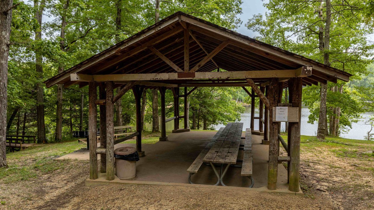 Wooden picnic shelter by a lake with picnic tables inside.
