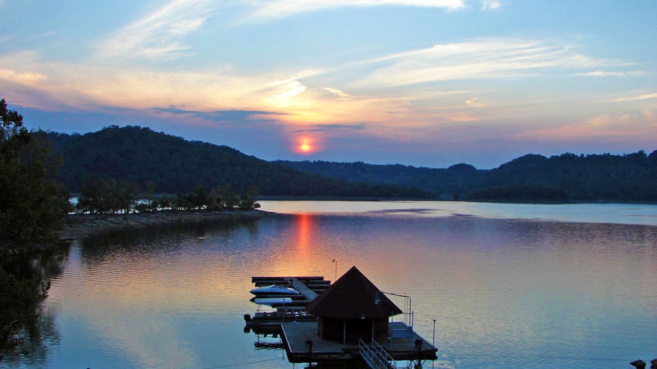 Sunset over a calm lake with a floating boathouse and forested hills.