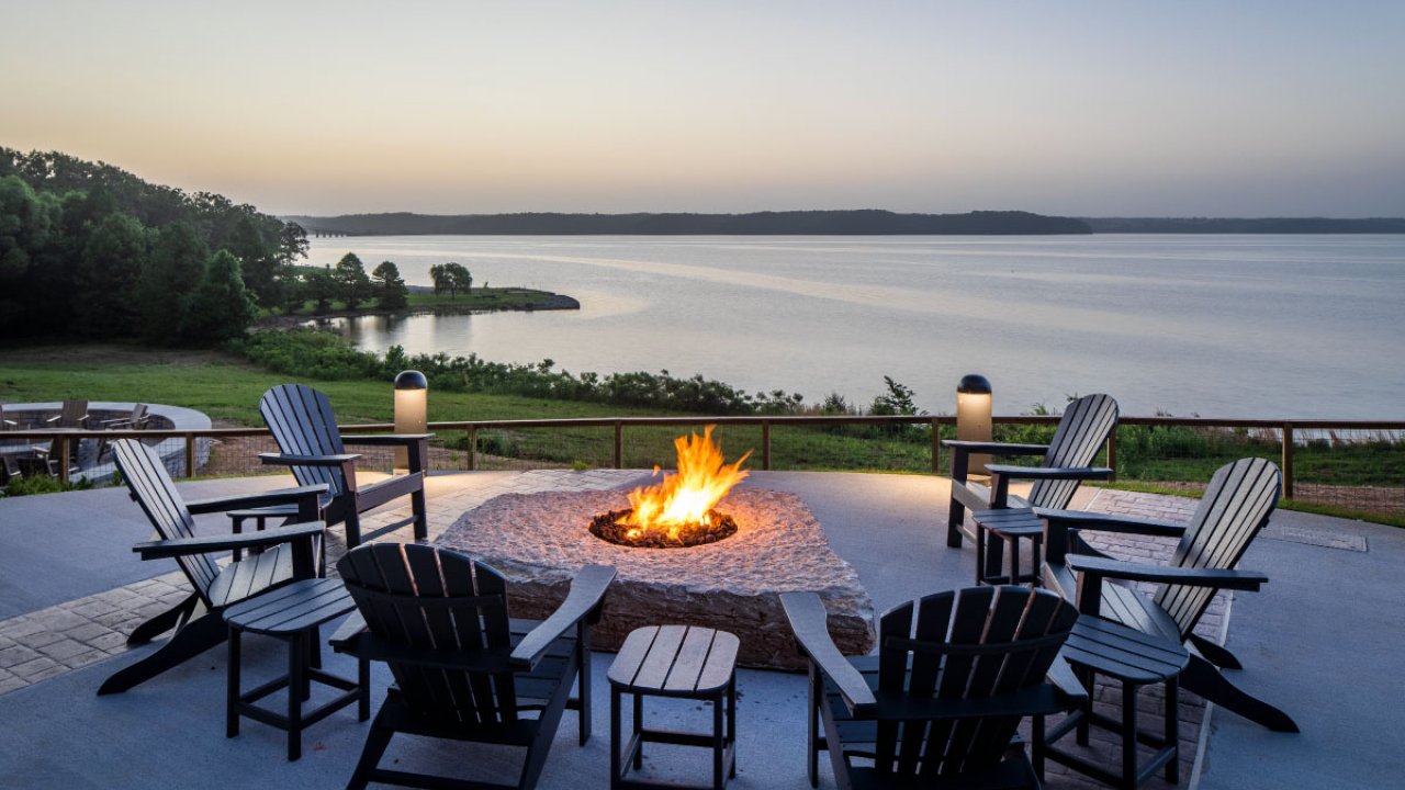 Outdoor patio with chairs around a fire pit overlooking a tranquil lake at dusk.
