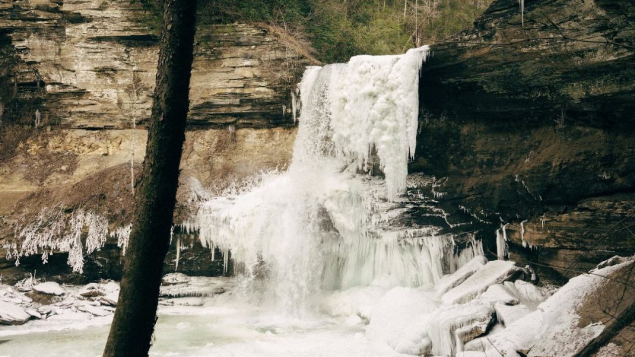 Partially frozen waterfall with icy cliffs and snow-covered ground.