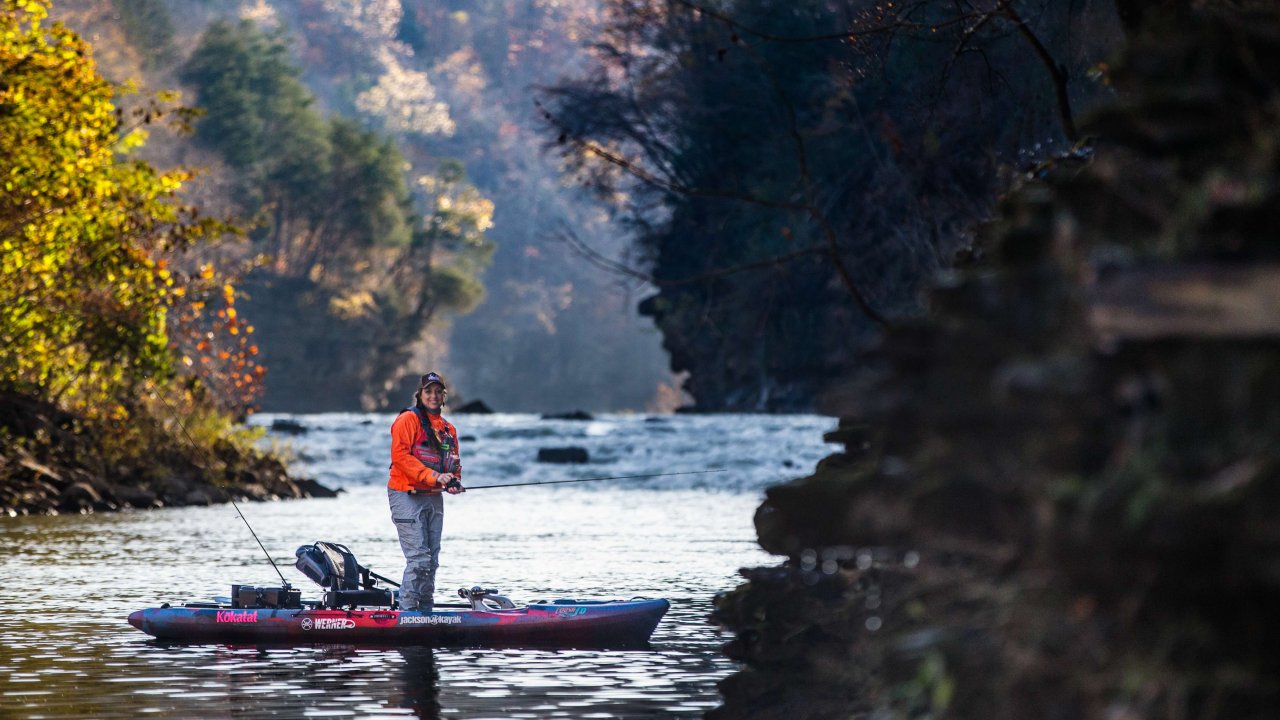 Person fishing from a kayak on a serene river surrounded by autumn trees.