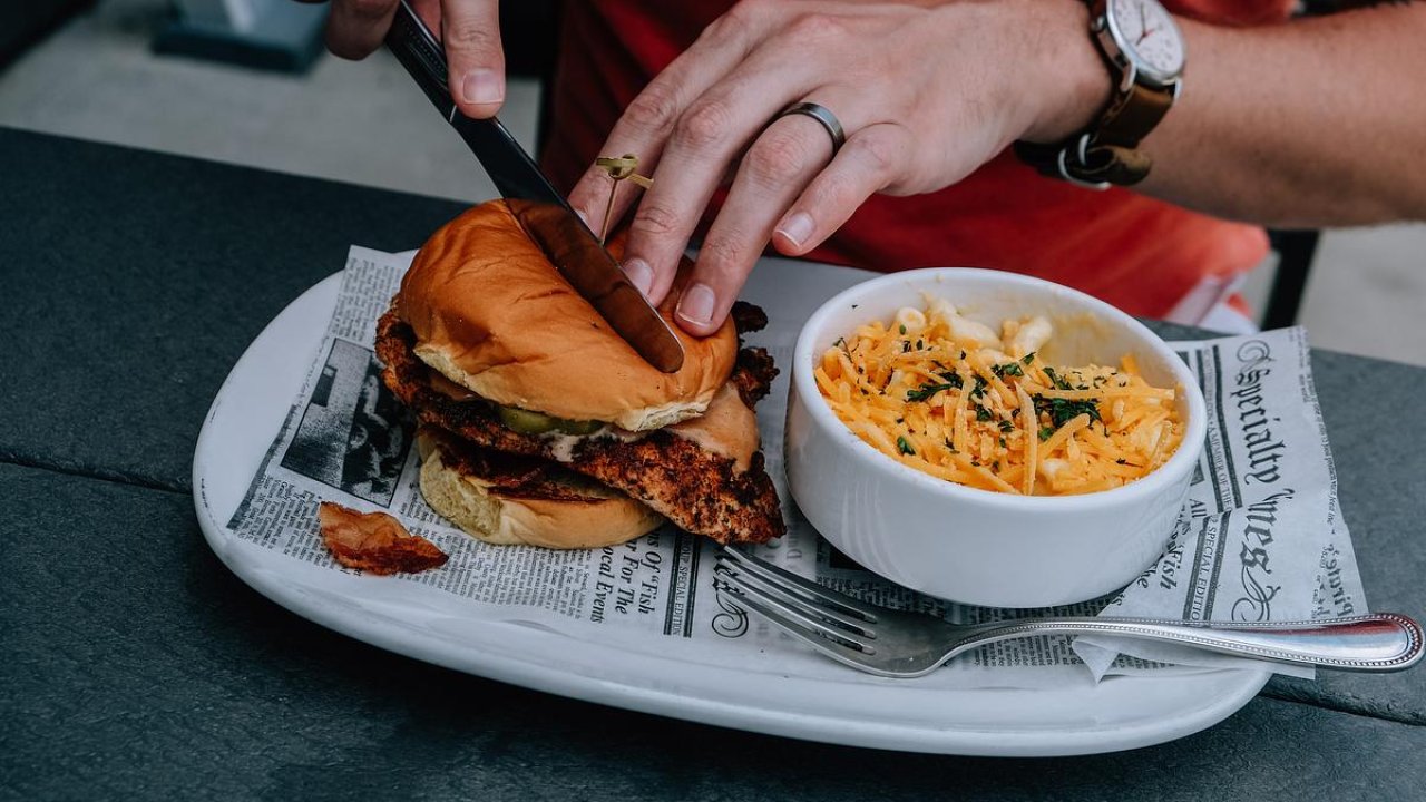 Person cutting a chicken sandwich, served with coleslaw on a newspaper-lined plate.