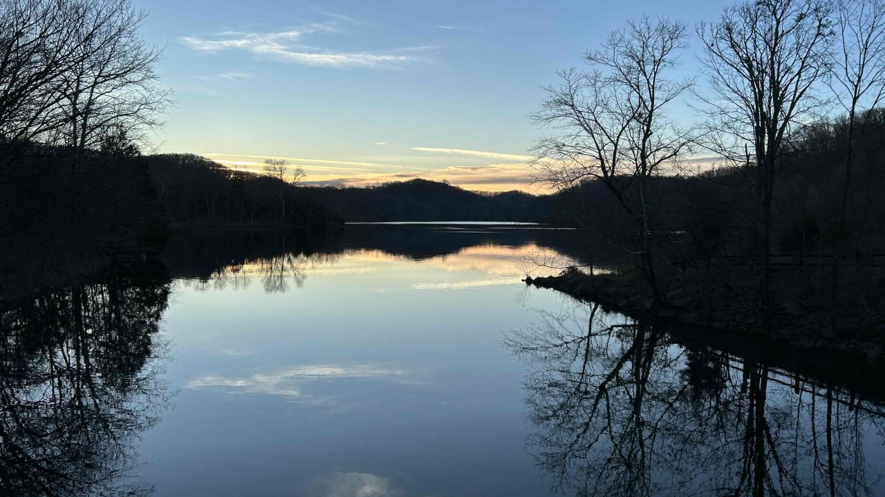 Tranquil sunset over a still lake with bare trees reflecting in the water.