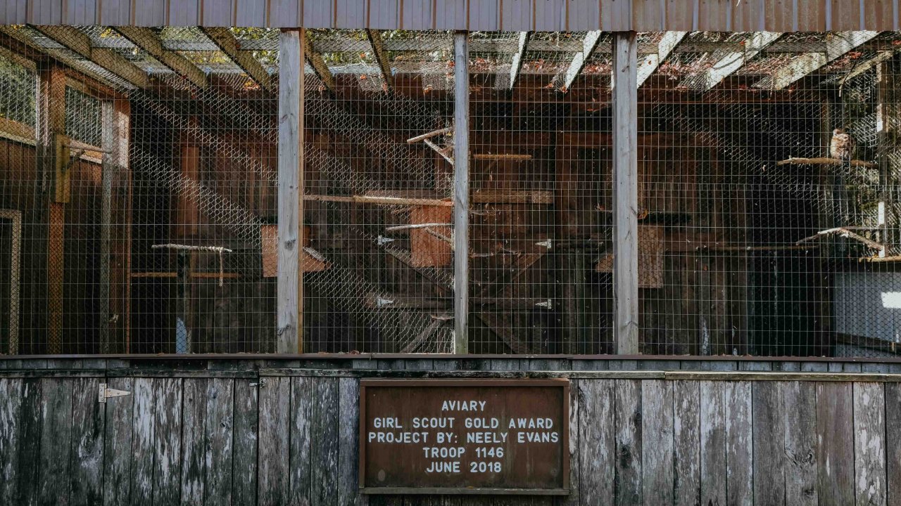 Rusty outdoor birdcage with a weathered wooden sign under a shaded roof.