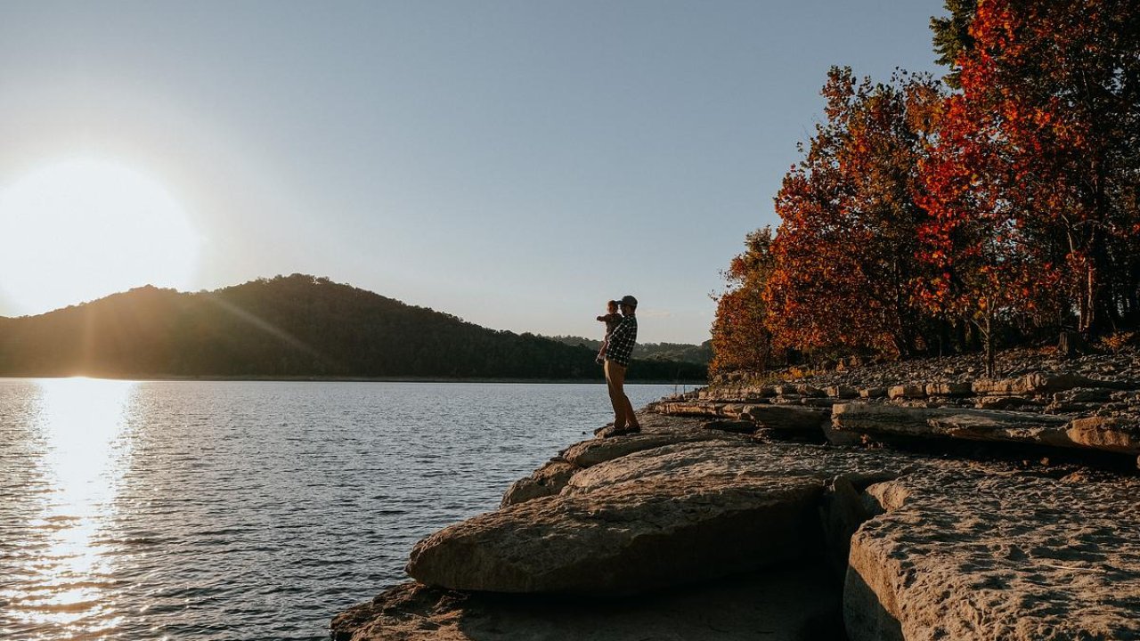 Person standing on a lakeside rock during autumn sunset.