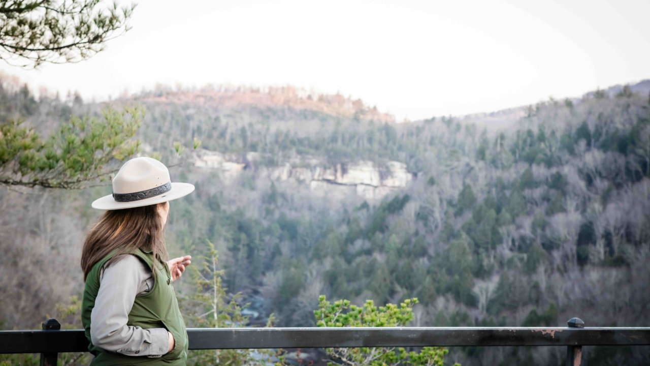 Woman in a hat admiring a mountainous landscape from a balcony.