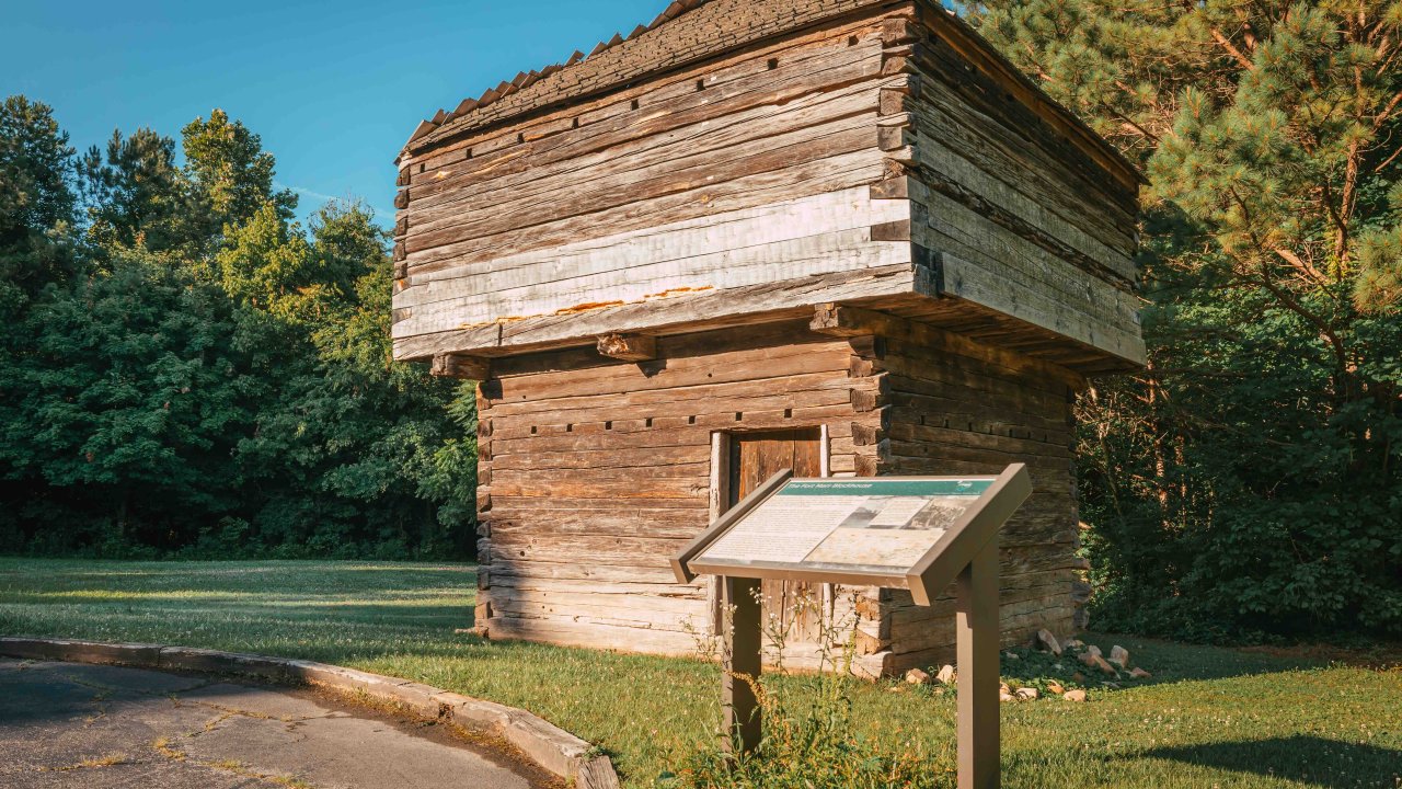 Historic wooden log cabin with an informational sign in a sunny park setting.