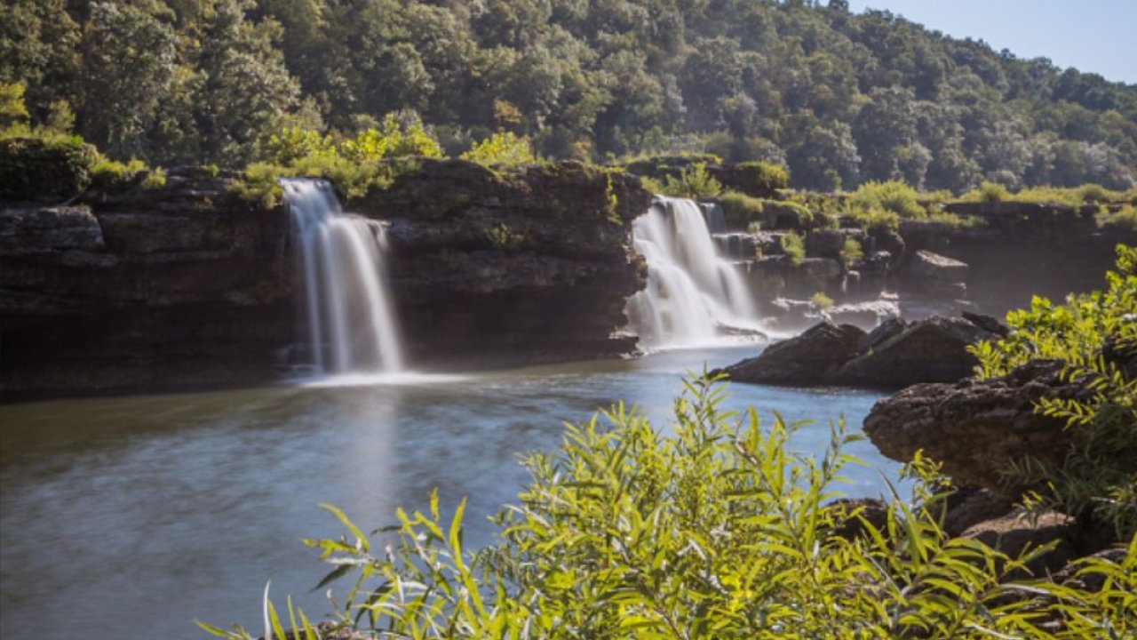 Waterfalls cascade into a calm river, surrounded by lush greenery and forest.