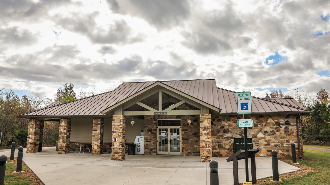 A brown, brick building with double doors stands in the middle of a concrete path under a cloudy sky. An accessible parking sign is visible to the right. 