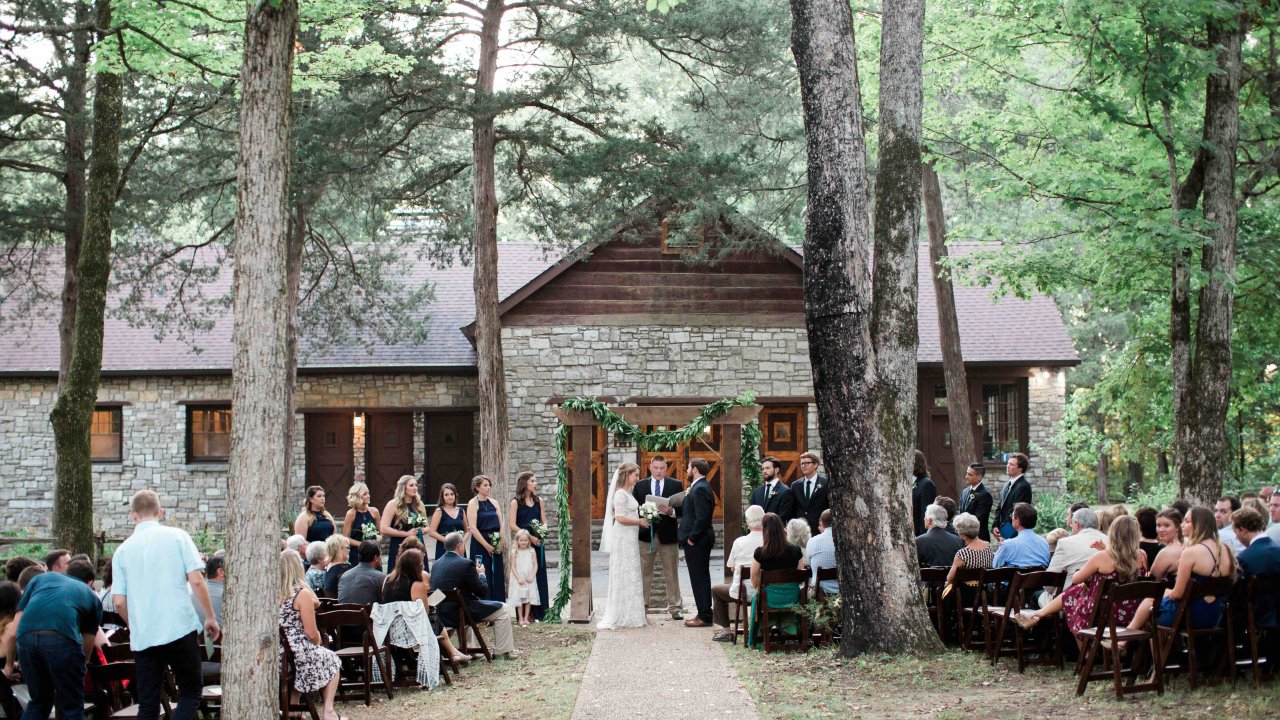 Outdoor wedding ceremony in a forest, guests seated along a path leading to a wooden cabin.