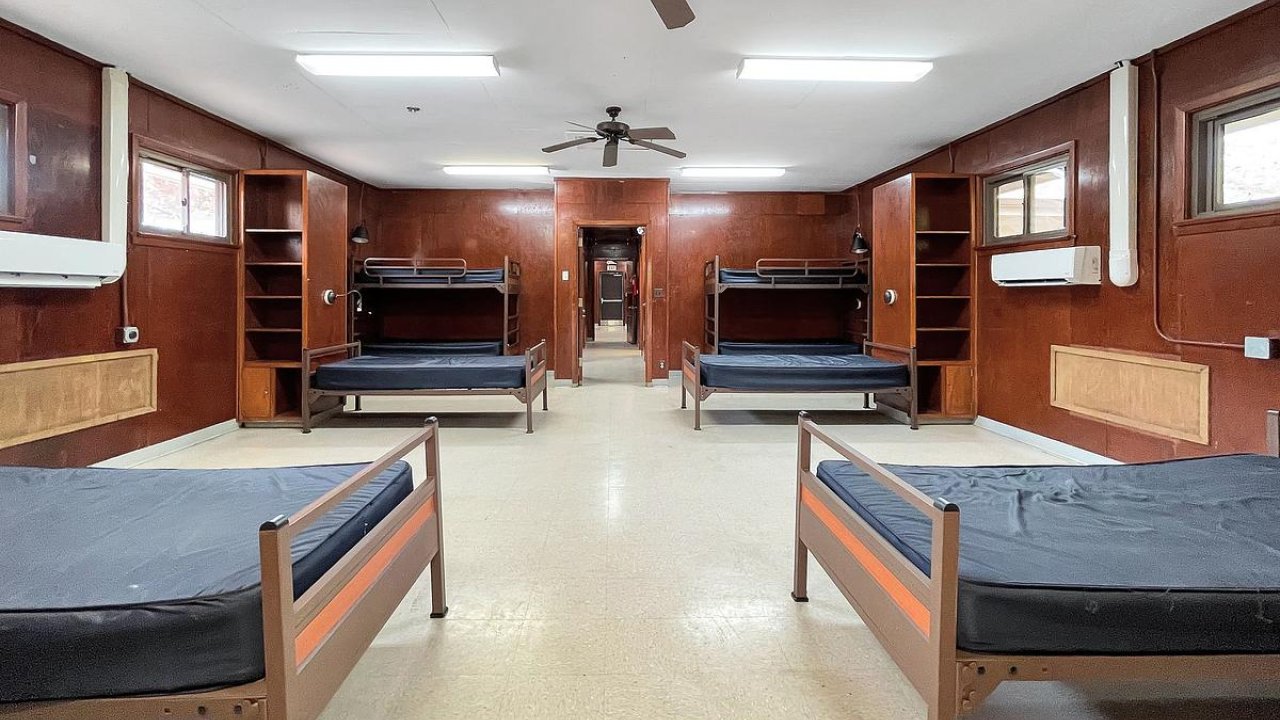 Empty dormitory room with twin beds, wooden shelves, and a ceiling fan.