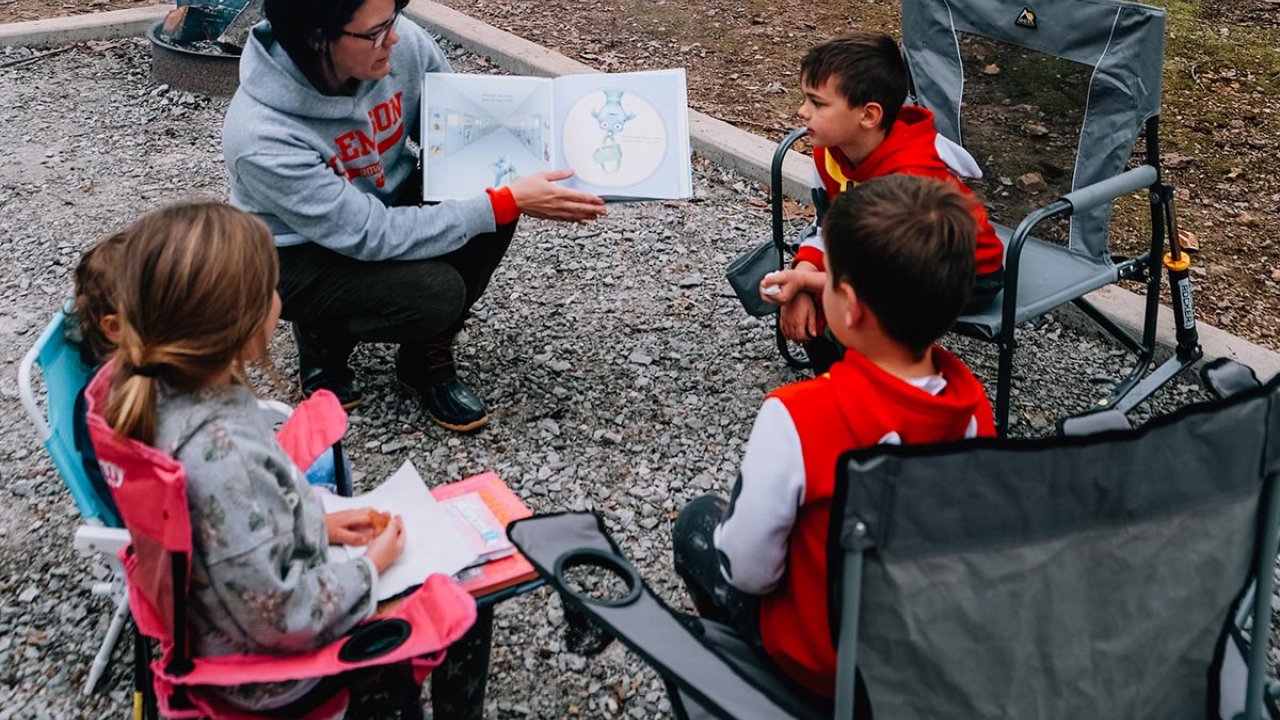 Instructor showing a book to three children sitting on camping chairs.