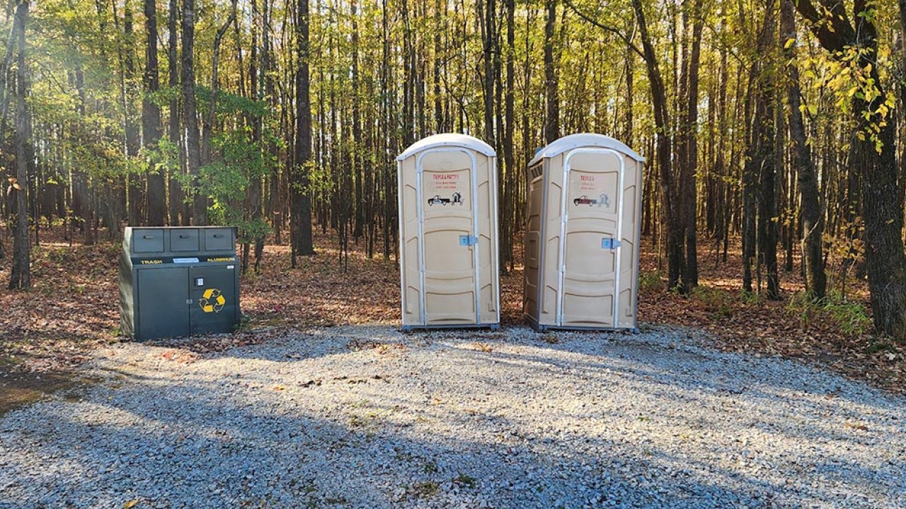 Two portable toilets on gravel, surrounded by trees in sunlight.