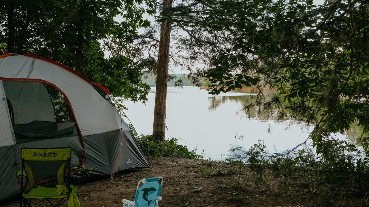 Camping setup by a lakeside with chairs and a tent under trees.