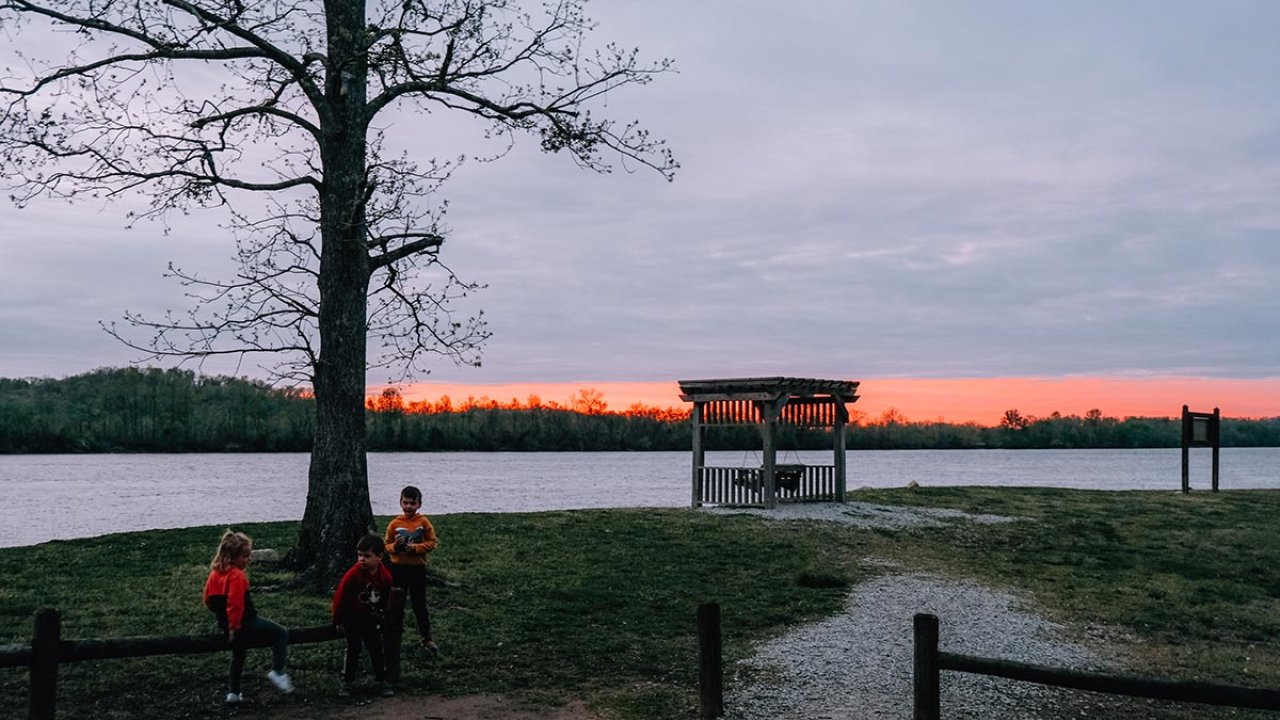 Sunset by a lake with three people sitting under a tree.