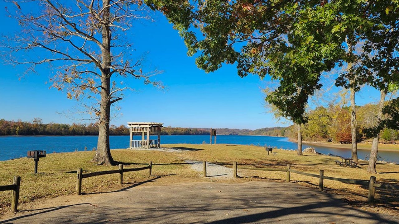 Pathway by a lake, lined with trees and a wooden shelter under a clear blue sky.