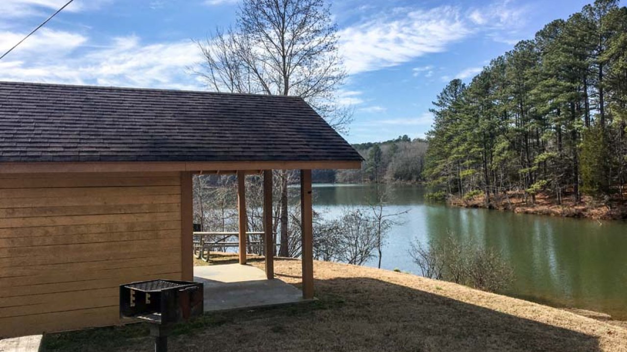 Cabin by a lake with trees, a small grill, and a clear blue sky.