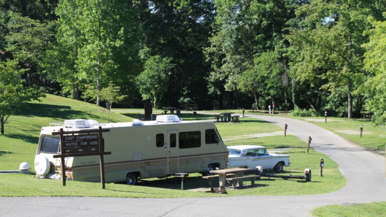 RV and vintage car parked in a lush, green campsite under sunny skies.