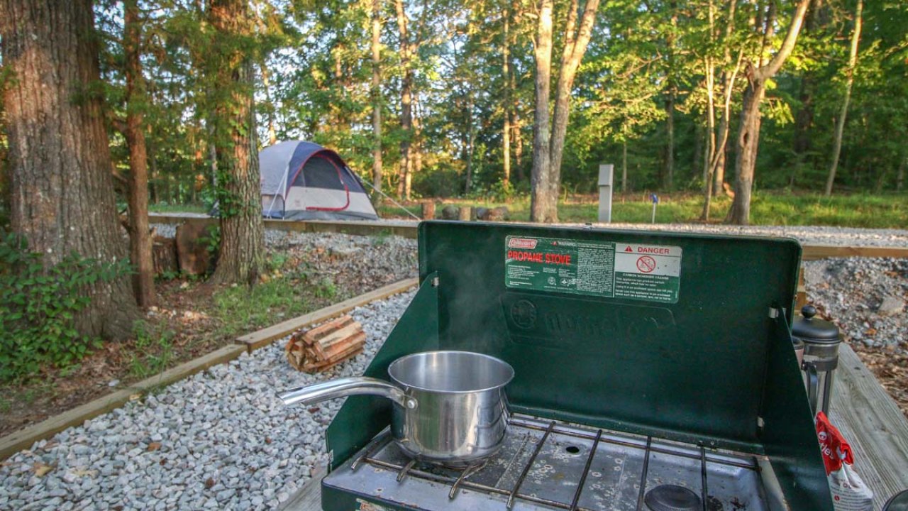 Camping stove with pot, tent in forest background.
