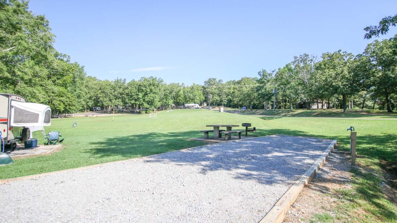 Sunny park with a gravel area, picnic table, and RV on grassy field.