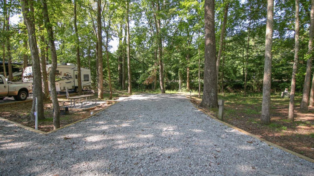Gravel path with trees and an RV in a wooded campsite.