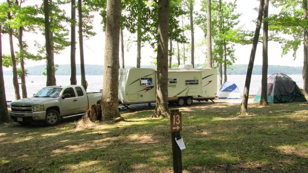 Truck and camper trailer parked among trees near a lake, with tents in the background.