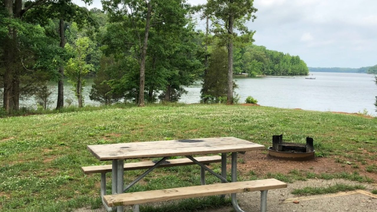 Picnic area with a table and fire pit by a tranquil lake surrounded by trees.