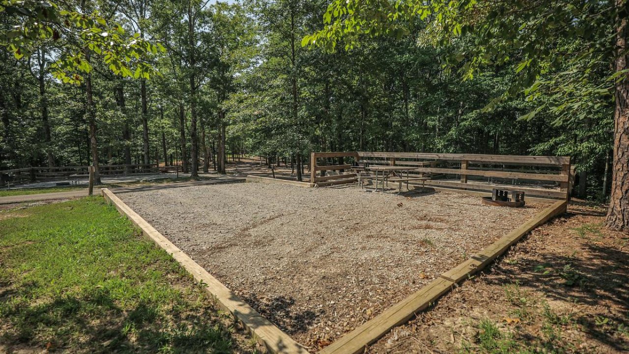 Empty horseshoe pit with surrounding fence in a forest clearing.