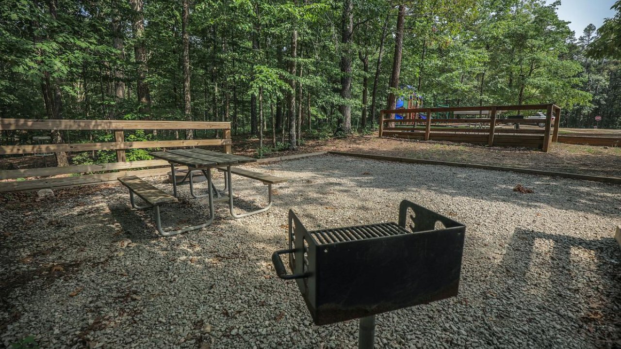 Empty campground site with a grill and picnic table, surrounded by trees.