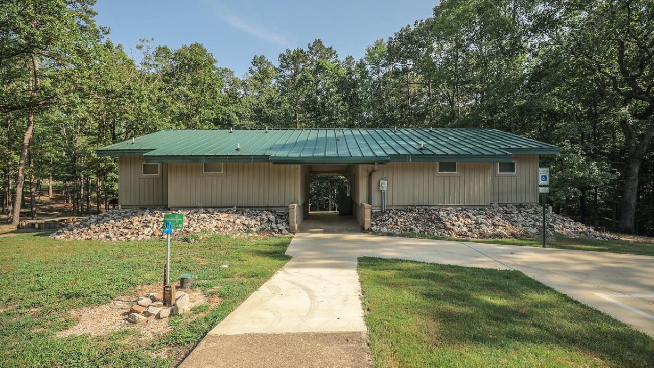 Single-story building with a green roof, surrounded by trees, viewed from a concrete pathway.