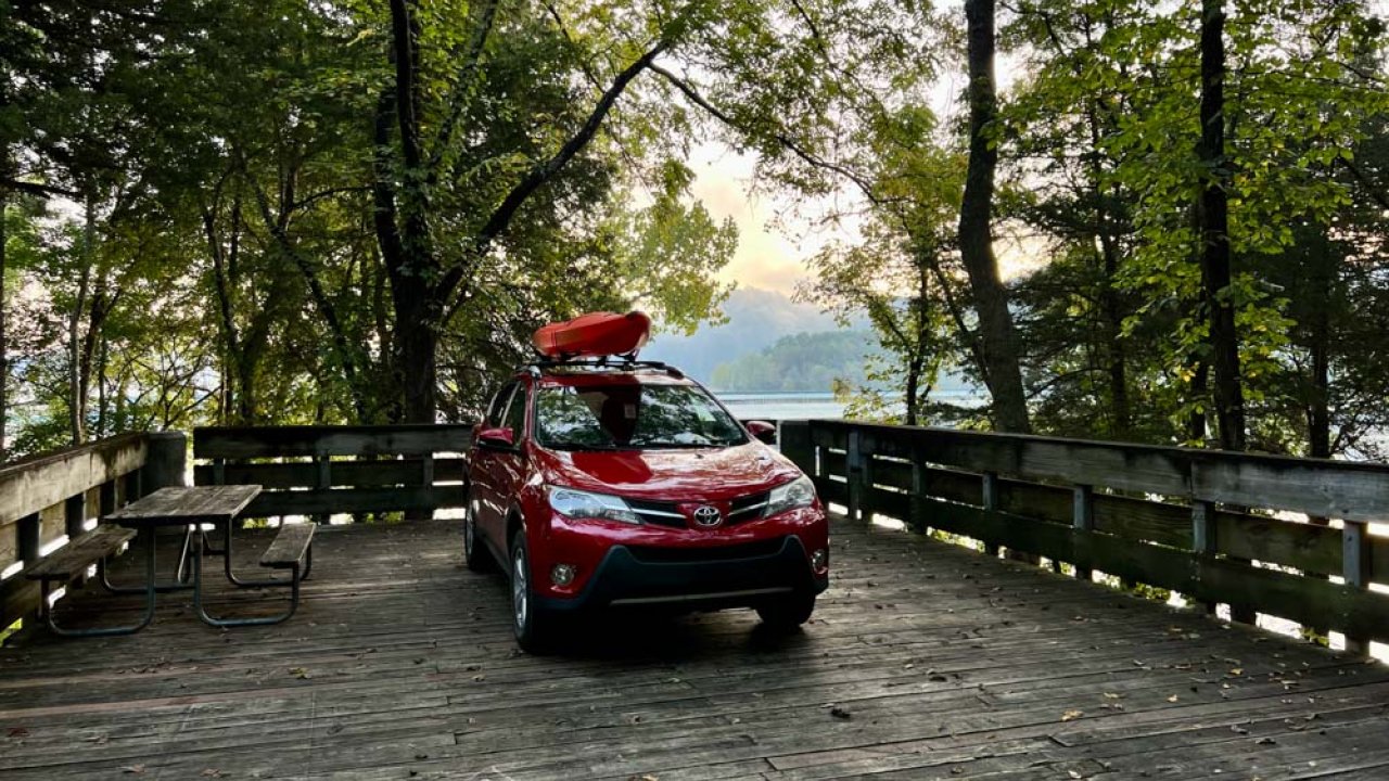 Red SUV with a kayak on top parked in a wooded lakeside area.