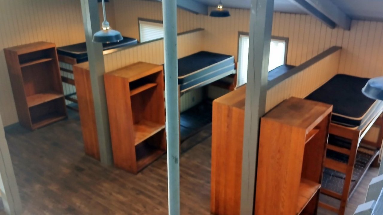 Hostel room with wooden bunk beds and shelves, lit by natural light.