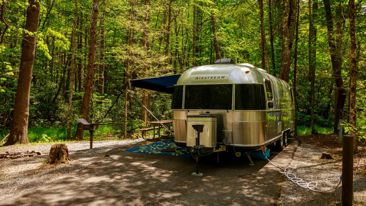 Silver trailer parked in a wooded campsite with lush green trees.
