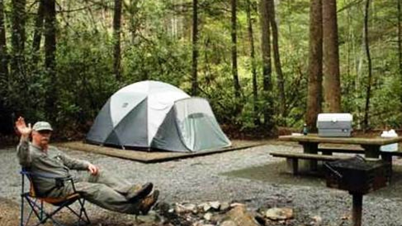 Man sitting near a tent in a forest campsite, waving, with a grill and picnic table nearby.