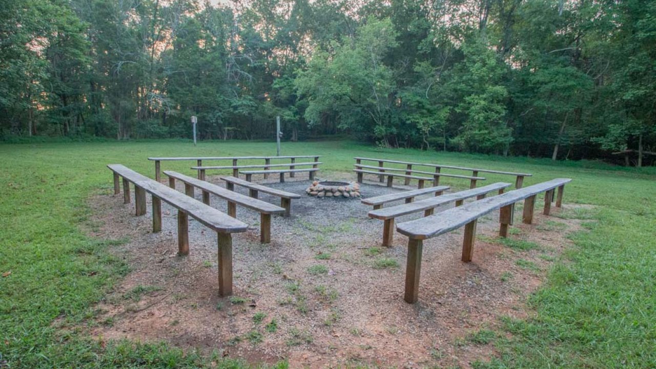Outdoor amphitheater with rows of wooden benches and central fire pit in a forest clearing.