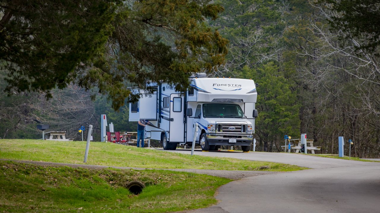 A motorhome parked at a paved RV campsite surrounded by trees, with picnic tables and utility hookups visible nearby.