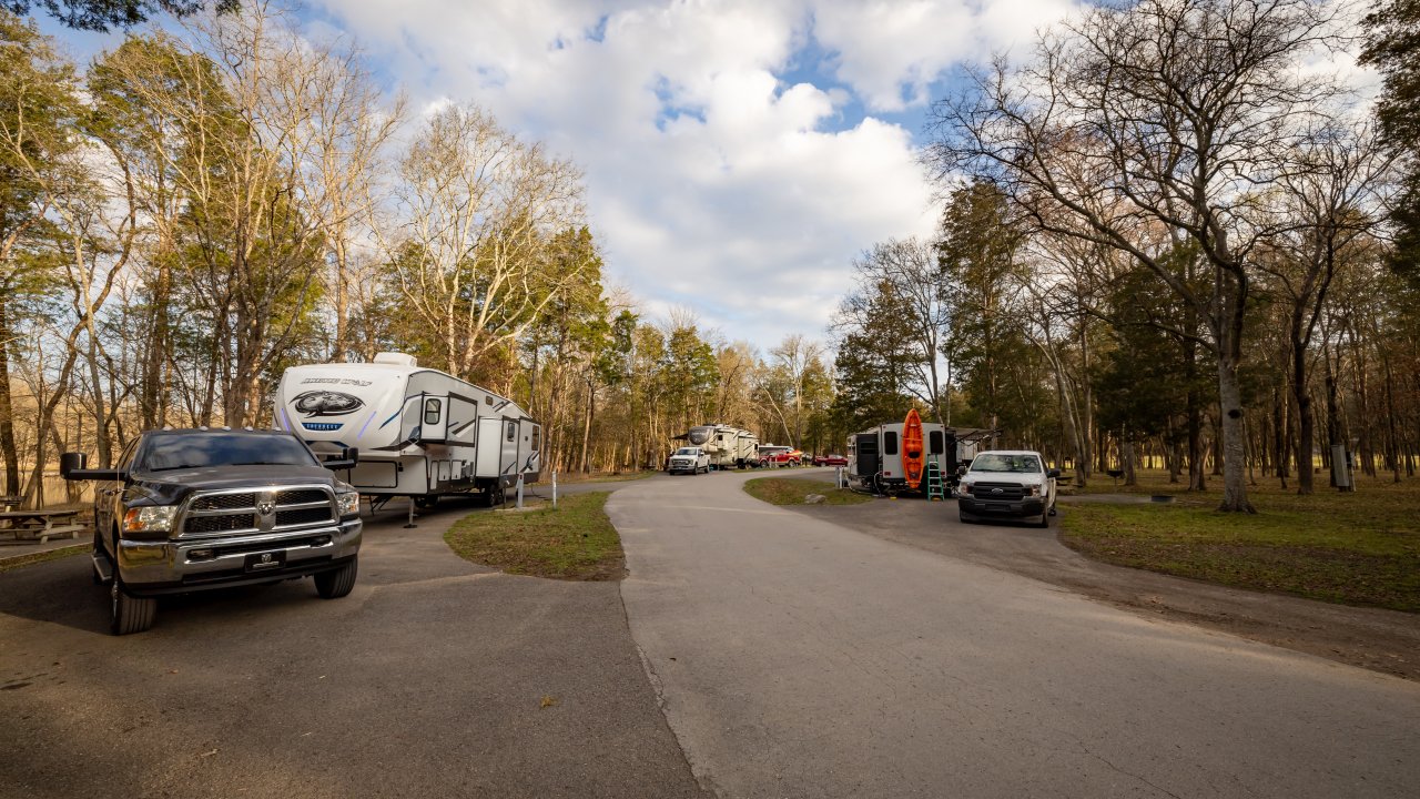 A paved campground road lined with RVs, trucks, and campsites set among tall trees on a clear day.
