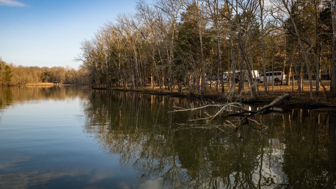 A calm lake reflecting trees along the shoreline, with RVs and campsites visible through the woods near the water’s edge.