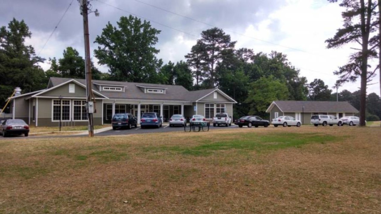 Large gray building with parked cars, surrounded by trees and grassy field. Cloudy sky.
