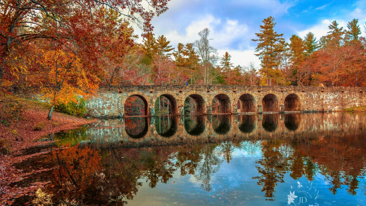Stone arch bridge spanning a calm body of water, surrounded by vibrant fall foliage in shades of red, orange, and gold. The bridge’s multiple arches are reflected clearly in the still water below, while trees with autumn leaves frame both sides of the scene under a partly cloudy blue sky.