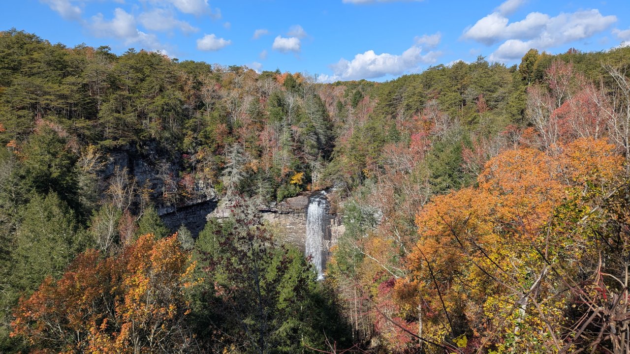 Waterfall cascading into a forested gorge surrounded by colorful fall foliage under a blue sky with scattered clouds.