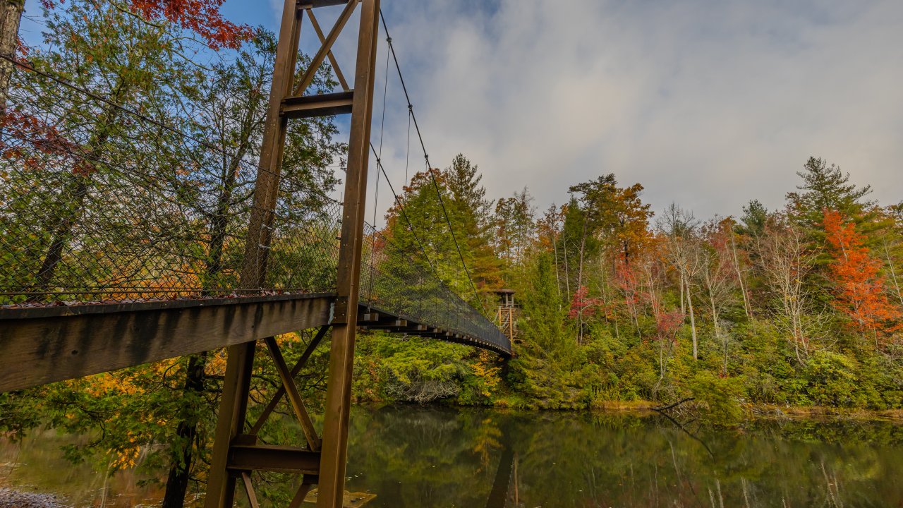 Suspension bridge over a calm lake surrounded by colorful autumn trees under a partly cloudy sky.