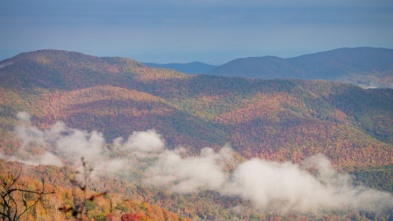 Scenic view of layered mountain ridges covered in vibrant fall foliage, with trees displaying shades of red, orange, yellow, and green. Low clouds or fog drift through the valleys between the hills, while a partly cloudy blue sky stretches above.
