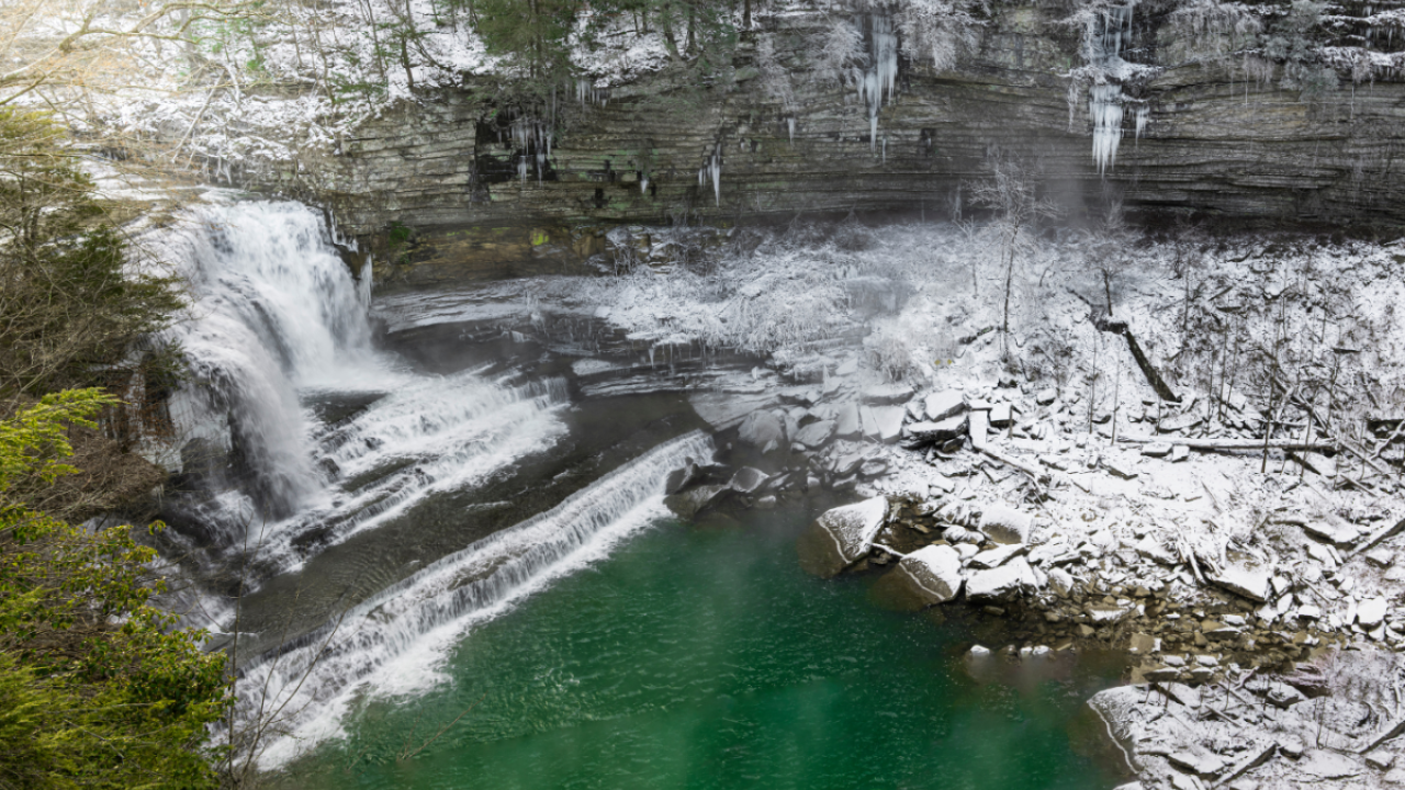 A large tiered waterfall spills into a blue pool surrounded by snow rocks and tall gorge walls