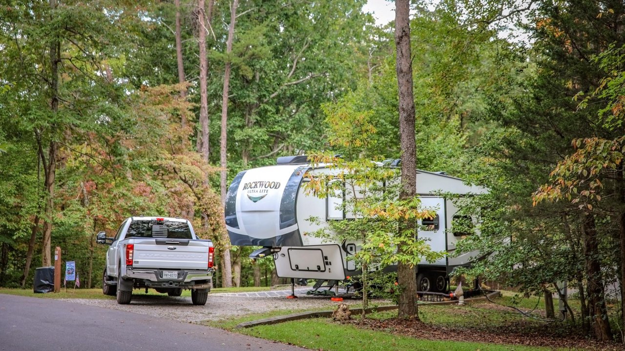 A white pickup truck is parked beside a large fifth-wheel RV labeled “Rockwood Ultra Lite,” set among trees with early fall foliage.